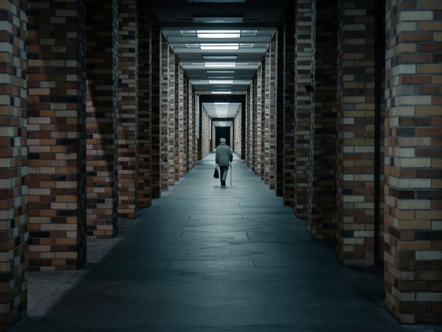 An older person walks alone down a long colonnade
